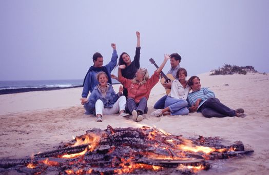 Group of people sitting on a beach at dusk with a campfire, singing songs with guitar