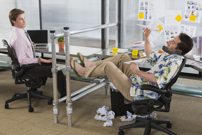 Two white men sitting in office, one wearing flip flops holding paper airplane wearing flower shirt, the other wearing pink shirt and tie